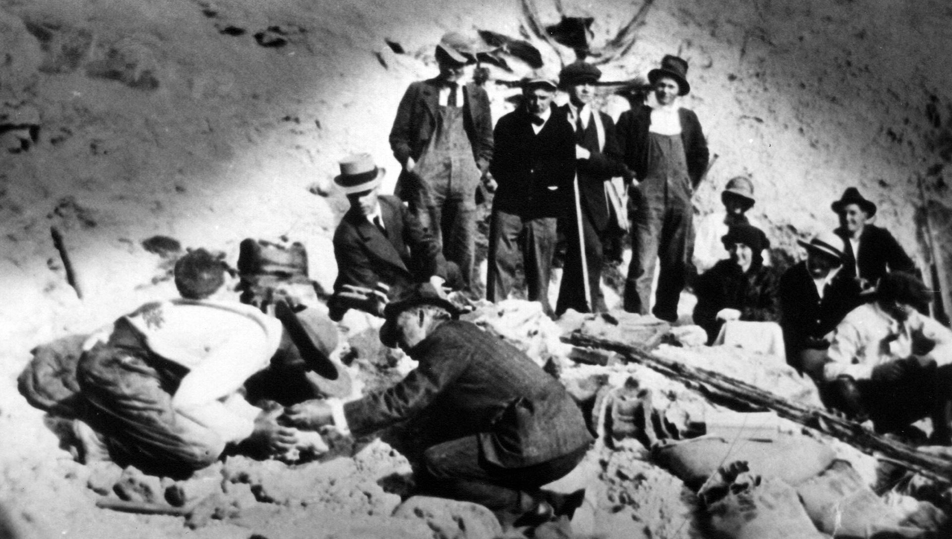 A grayscale photo of an unidentified group of people watching two men excavating a dinosaur bone.