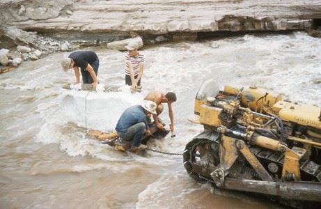 Color photo of jacketed dinosaur fossils on a wooden raft in a creek. Circa 1954.