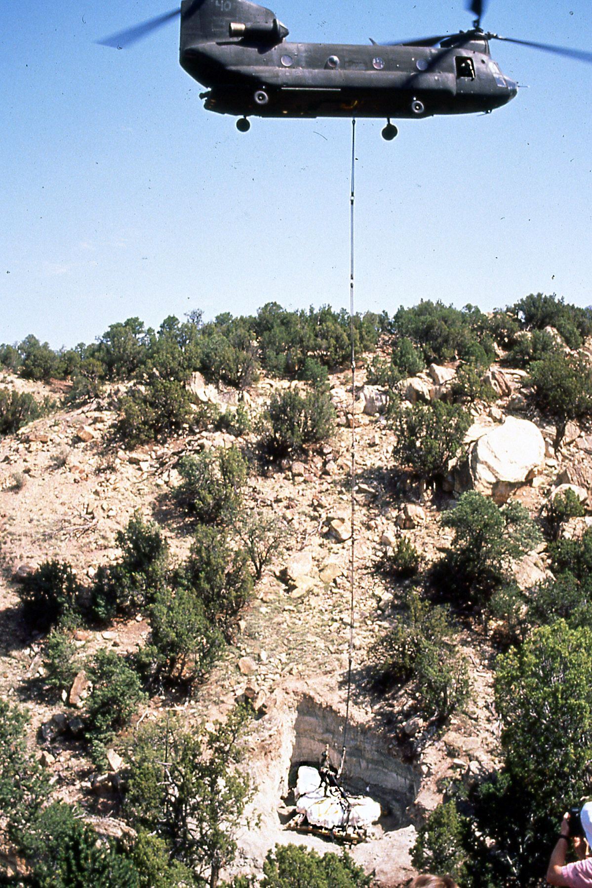 Color photo of Chinook helicopter hovering over dinosaur fossil quarry with stegosaurus attached.