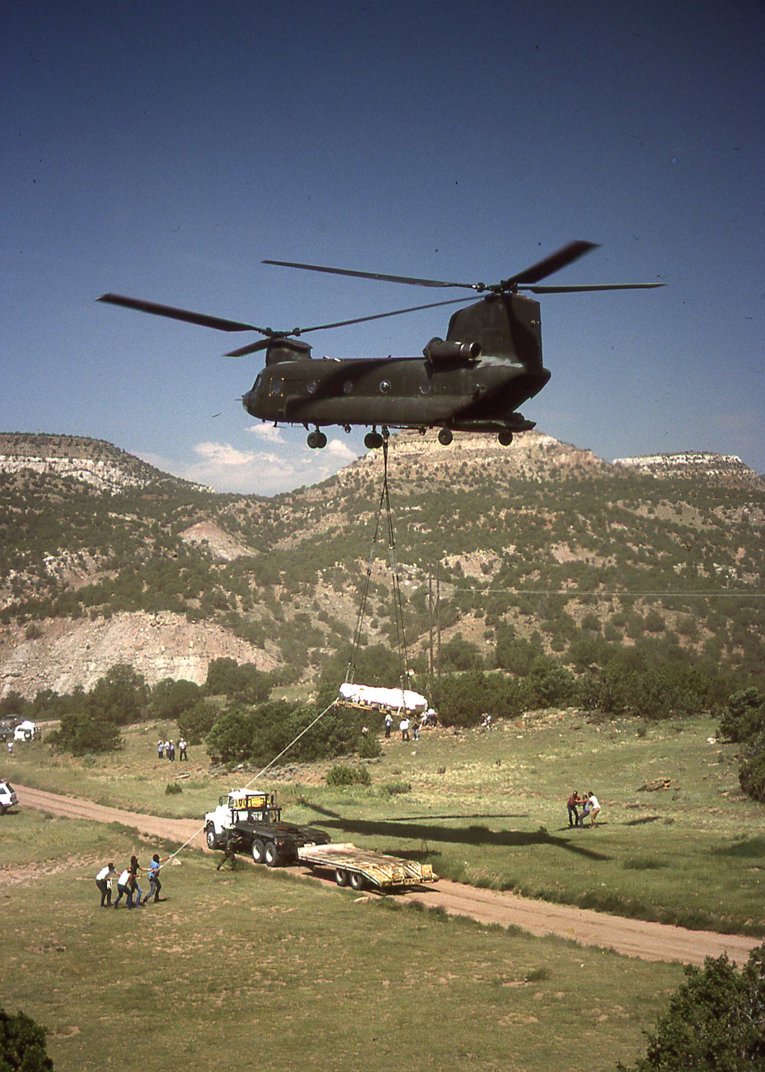 Color photo of Chinook helicopter flying over truck preparing to unload stegosaurus fossil.